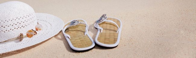 Strandutensilien: weiße Strandhaube, Flip-Flops mit Schmucksteinen, Sand.