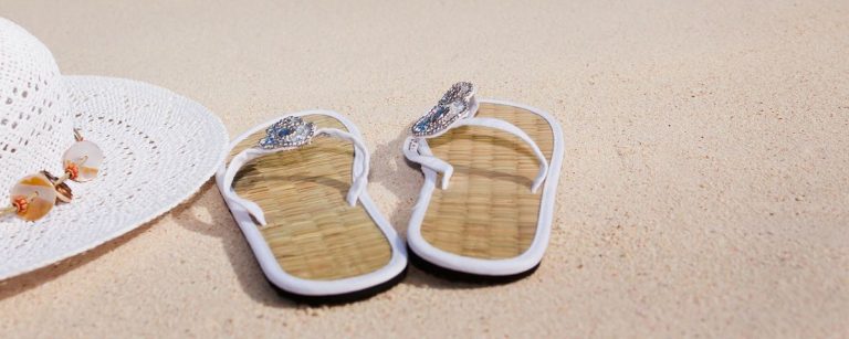 Strandutensilien: weiße Strandhaube, Flip-Flops mit Schmucksteinen, Sand.