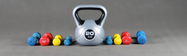 Ein großer silberner Kettlebell mit 20 kg Gewicht, umgeben von bunten Kurzhanteln in Rot, Gelb und Blau.