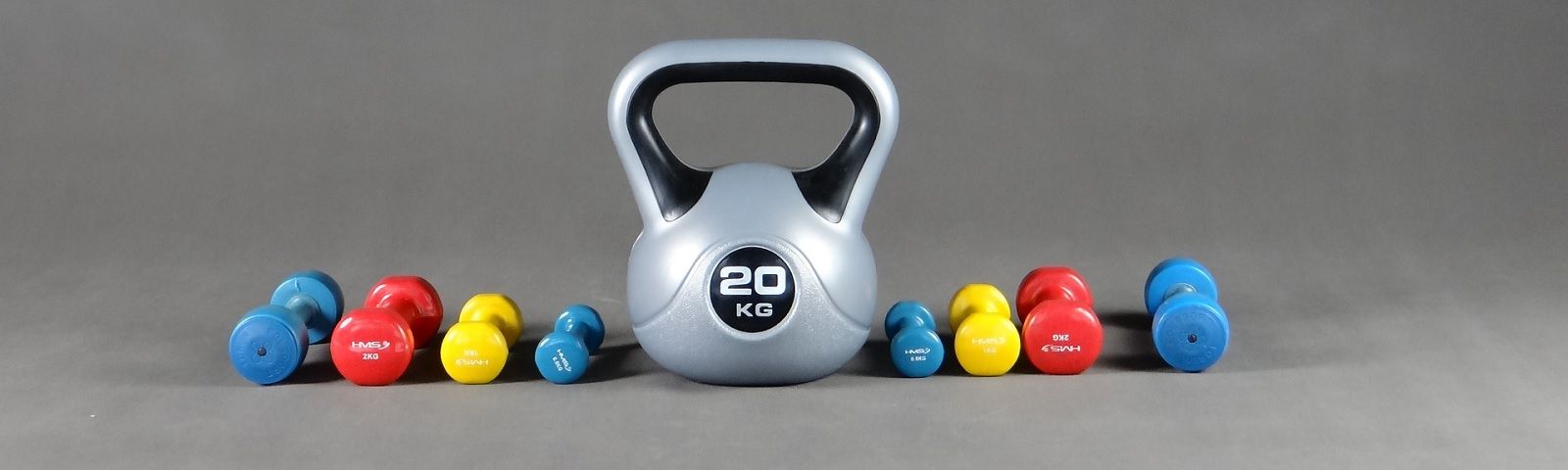 BF-1712-fitnesstrends-1600×480 Ein großer silberner Kettlebell mit 20 kg Gewicht, umgeben von bunten Kurzhanteln in Rot, Gelb und Blau.