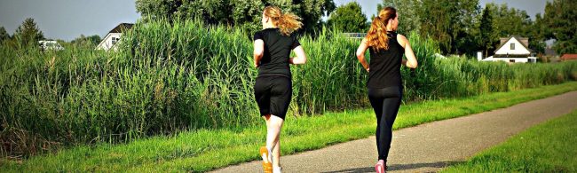 BF-bewegingsgebrek-1600×480 Zwei Frauen joggen auf einem Feldweg, umgeben von üppigem Grün, für ein Fitness- und Ernährungsunternehmen.