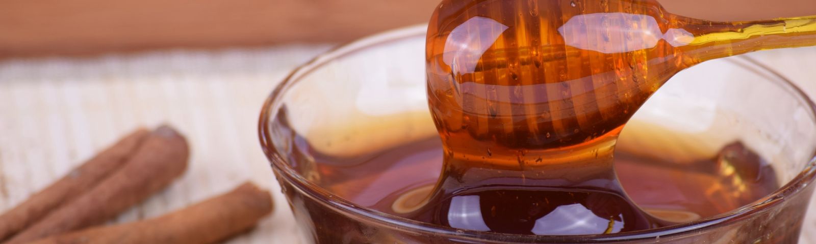 BF-honing-en-kaneel-1600×480 Goldener Honig fließt von einem Löffel in eine Schüssel, daneben Zimtstangen – perfekt für Fitness-Rezepte.