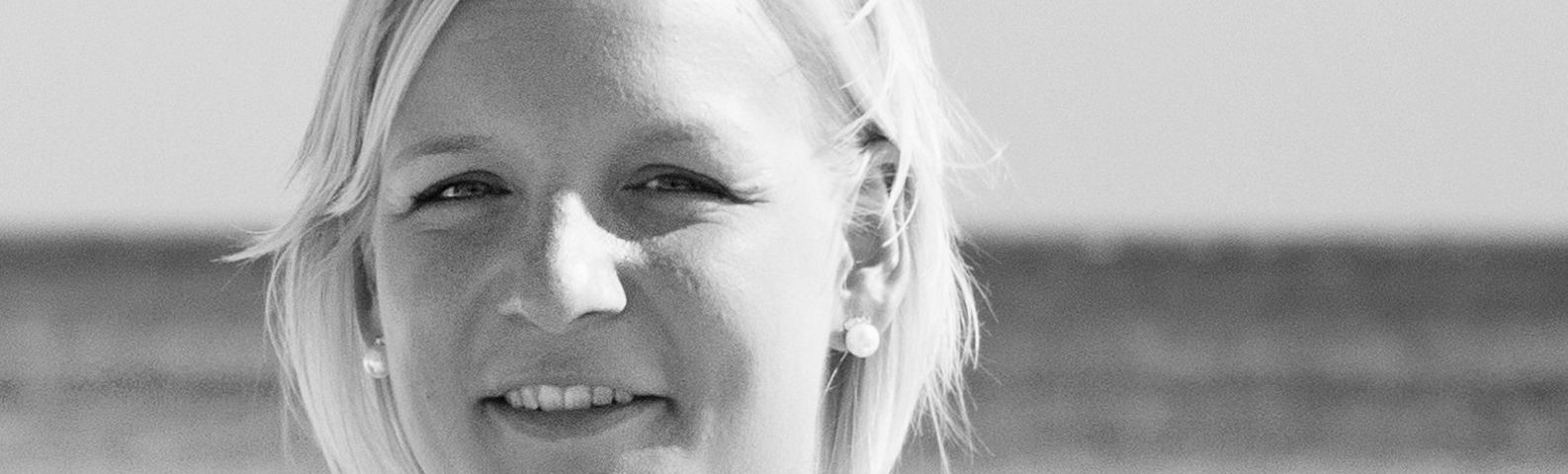 IMG-8-2-1 Frau mit blonden Haaren und Ohrringen lächelt in die Kamera, Porträt vor unscharfem Hintergrund.