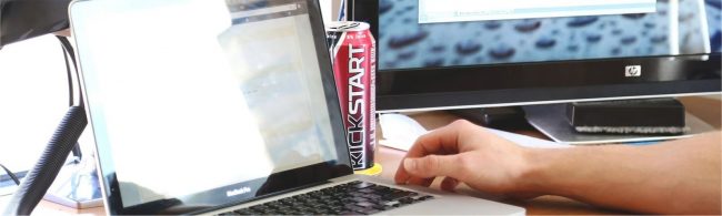 Ein Mann tippt auf seinem Laptop, neben ihm eine Kickstart-Dose und ein HP-Monitor mit Regentropfen.