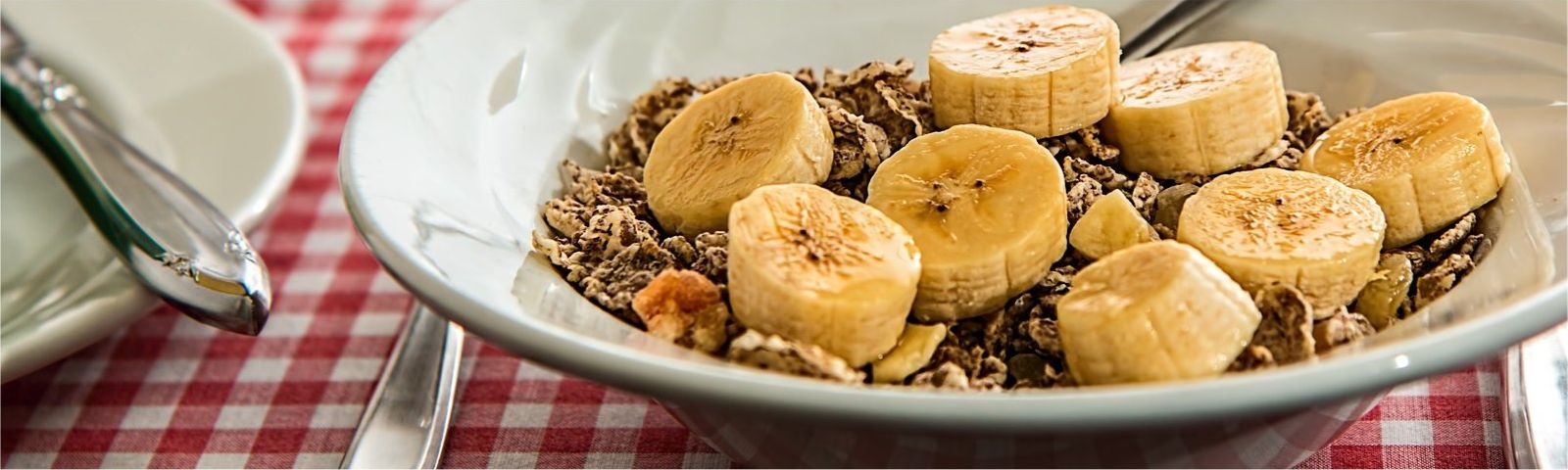 maart-vezels-groot_1 Müsli mit Bananenscheiben und einem Löffel auf karierter Tischdecke, perfekt für Fitness und gesunde Ernährung.