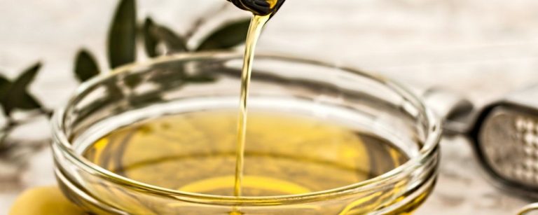Olivenöl fließt in eine Schüssel, mit Olivenzweigen und Knoblauchpresse im Hintergrund.