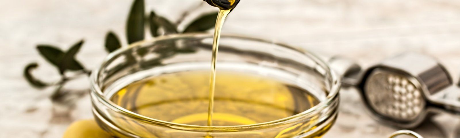 olive-oil-968657-1920_1 Olivenöl fließt in eine Schüssel, mit Olivenzweigen und Knoblauchpresse im Hintergrund.