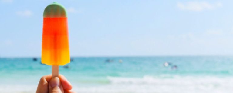 Ein handgehaltener Eis am Stiel vor einem hellblauen Himmel und Meer, das einen gesunden Snack für heiße Tage symbolisiert.