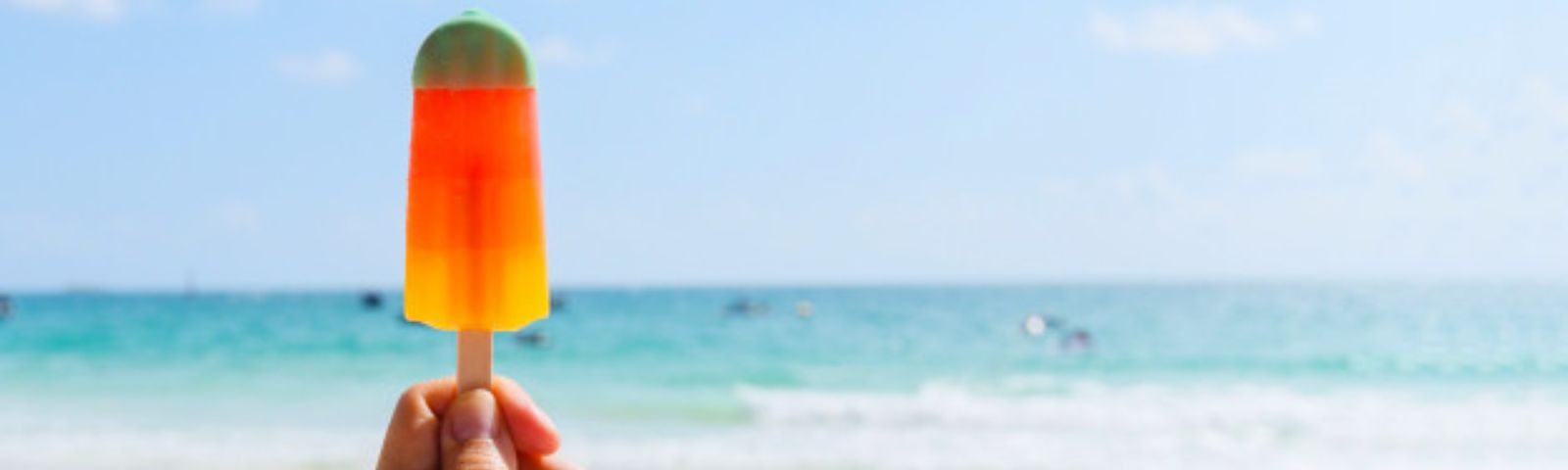 Ein handgehaltener Eis am Stiel vor einem hellblauen Himmel und Meer, das einen gesunden Snack für heiße Tage symbolisiert.