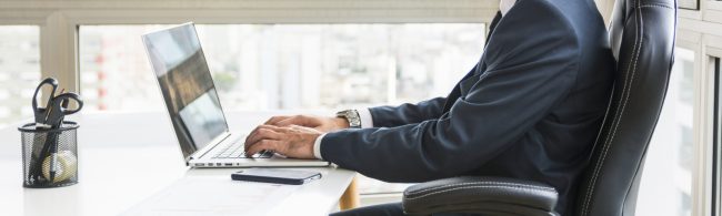 Mann im Anzug tippt auf Laptop, neben einer Schere im Behälter.