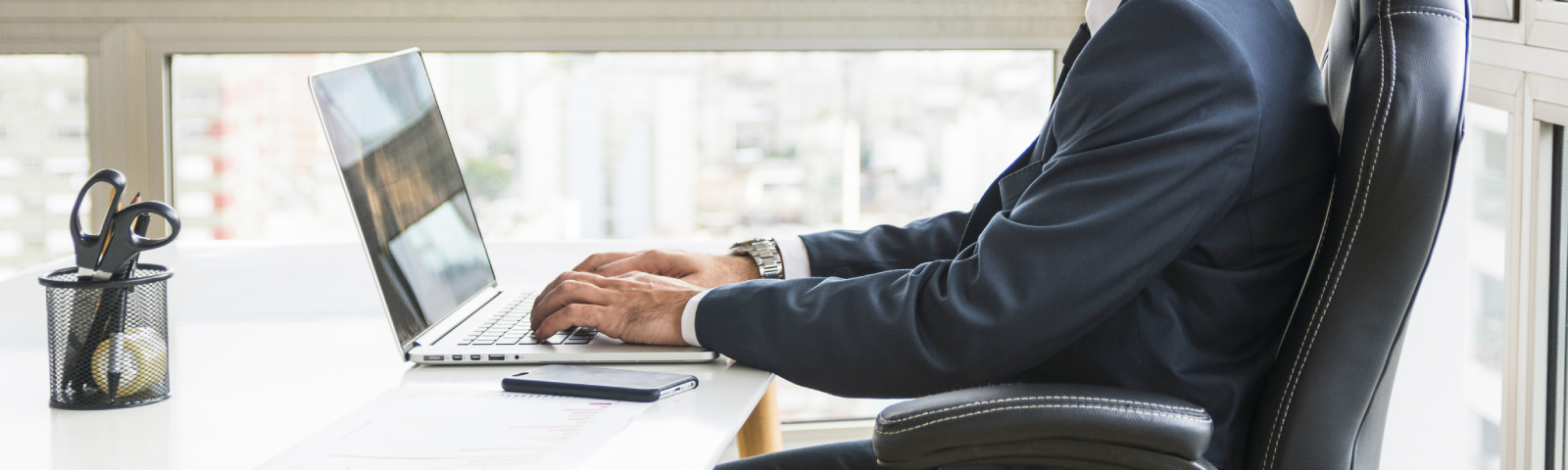 Mann im Anzug tippt auf Laptop, neben einer Schere im Behälter.