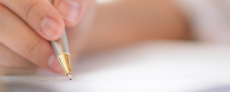 Hand mit Stift schreibt auf einem Dokument, im Hintergrund ein helles, natürliches Licht.