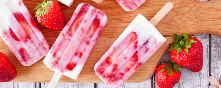 Vegane Erdbeer-Joghurt-Eisriegel auf Holzbrett mit frischen Erdbeeren.