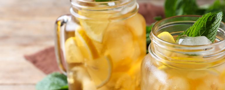 Pitcher and glasses of homemade iced tea with ice, citrus slices, fresh herbs and a tray of fruit on a sunny table.