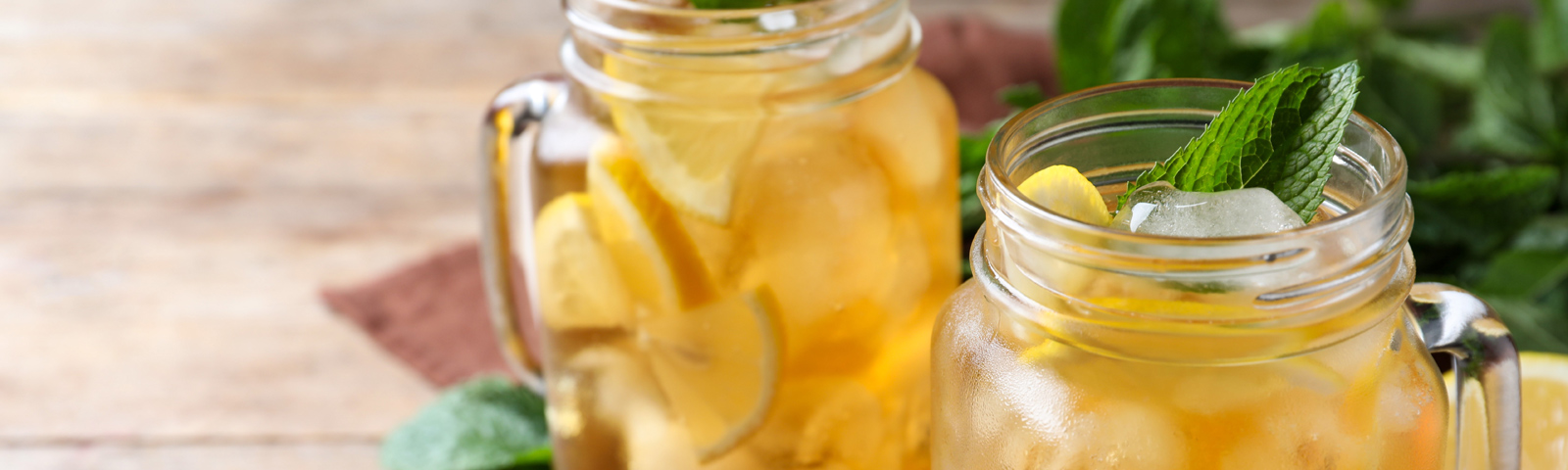 Pitcher and glasses of homemade iced tea with ice, citrus slices, fresh herbs and a tray of fruit on a sunny table.