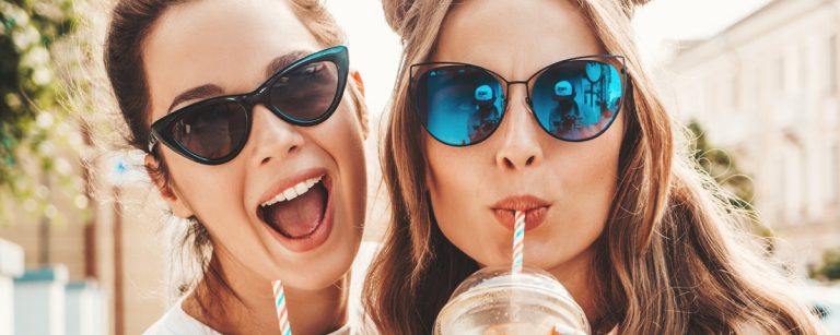Zwei junge Frauen in Sonnenbrillen trinken gesunde Smoothies im Freien.