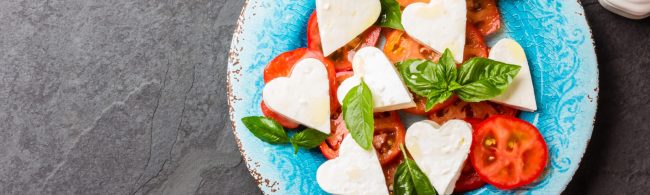 A plated three-course meal for Valentine’s Day featuring heart-shaped mozzarella Caprese salad, herbed grilled chicken with lemon, and chocolate-covered fruit treats.