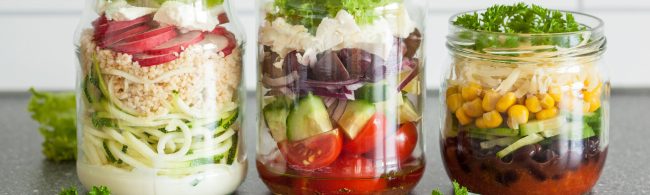 A colorful layered Mexican salad in a glass jar, with beans, corn, tomatoes, avocado, carrots, red onion and lime dressing neatly visible.