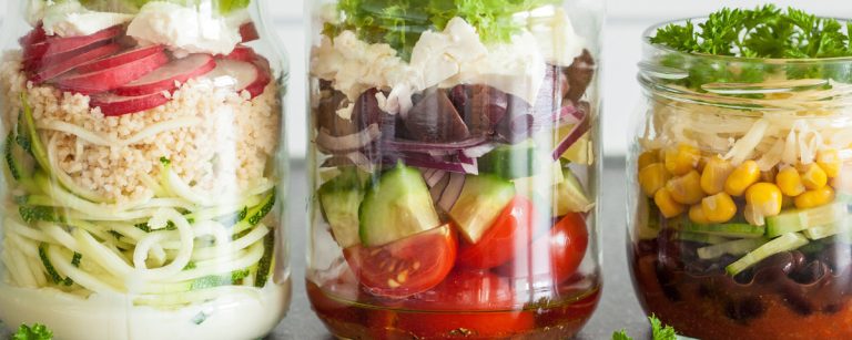 A colorful layered Mexican salad in a glass jar, with beans, corn, tomatoes, avocado, carrots, red onion and lime dressing neatly visible.