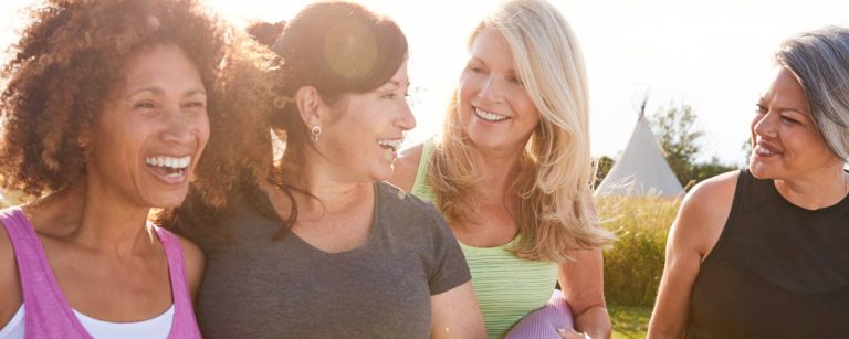 Two friends exercising together outdoors, smiling and doing bodyweight squats side by side in a sunny park.
