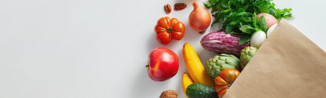 aper grocery bag with assorted seasonal produce—tomatoes, apple, bananas, eggplant, cucumber, artichokes, onion, fresh herbs and nuts—on a light background