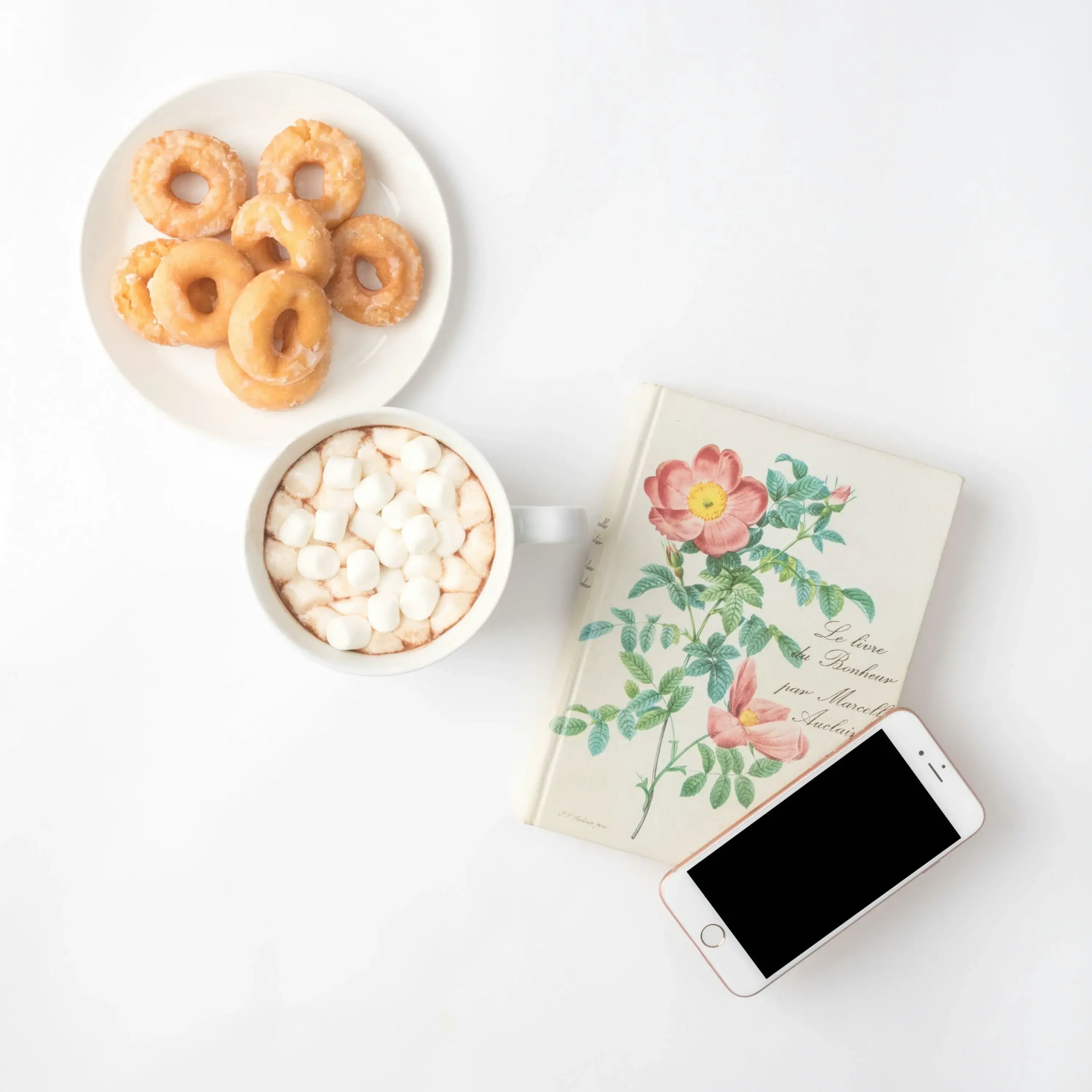 personalisierter-ernaehrungsplan-guide Draufsicht auf eine gemütliche Szene mit glasierten Donuts, Kakao mit Marshmallows, einem floralen Buch und einem Smartphone auf weißem Hintergrund. Dieses Bild kann einen Moment der Entspannung und des Genusses symbolisieren, um die Wichtigkeit von **Balance** und realistischer Integration von Leckereien in einen **gesunden Lebensstil** und eine **Fitness-Ernährung** zu unterstreichen.
